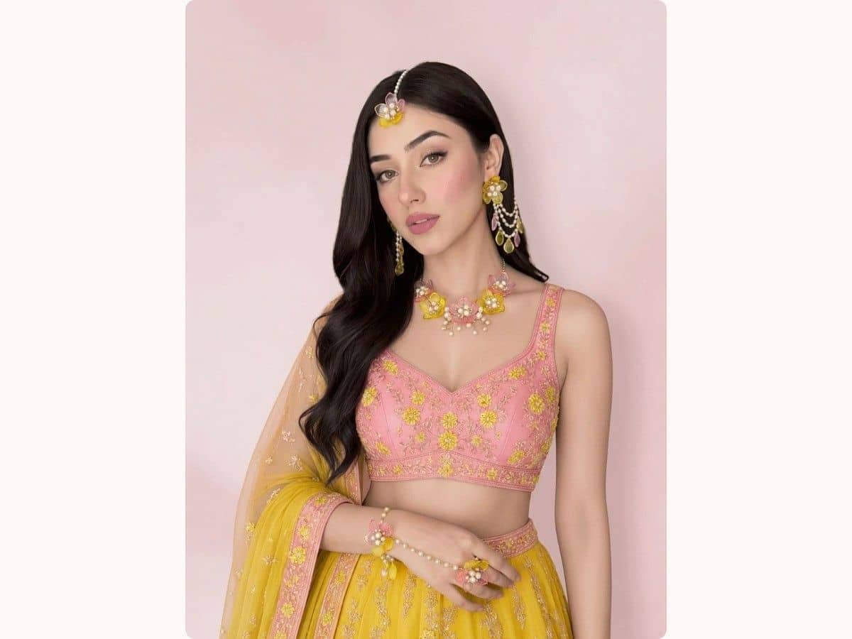 A bride in a yellow lehenga wearing a yellow and pink resin-flower maang tikka, earrings, necklace and haathphool