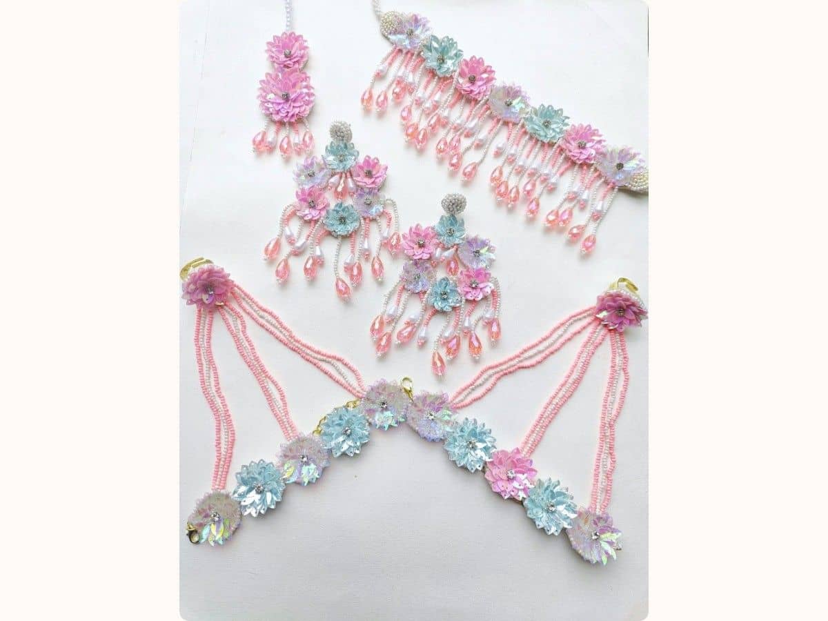Pastel pink and blue flower set laid flat: necklace with pink crystal drops, earrings, maang tikka and hand pieces